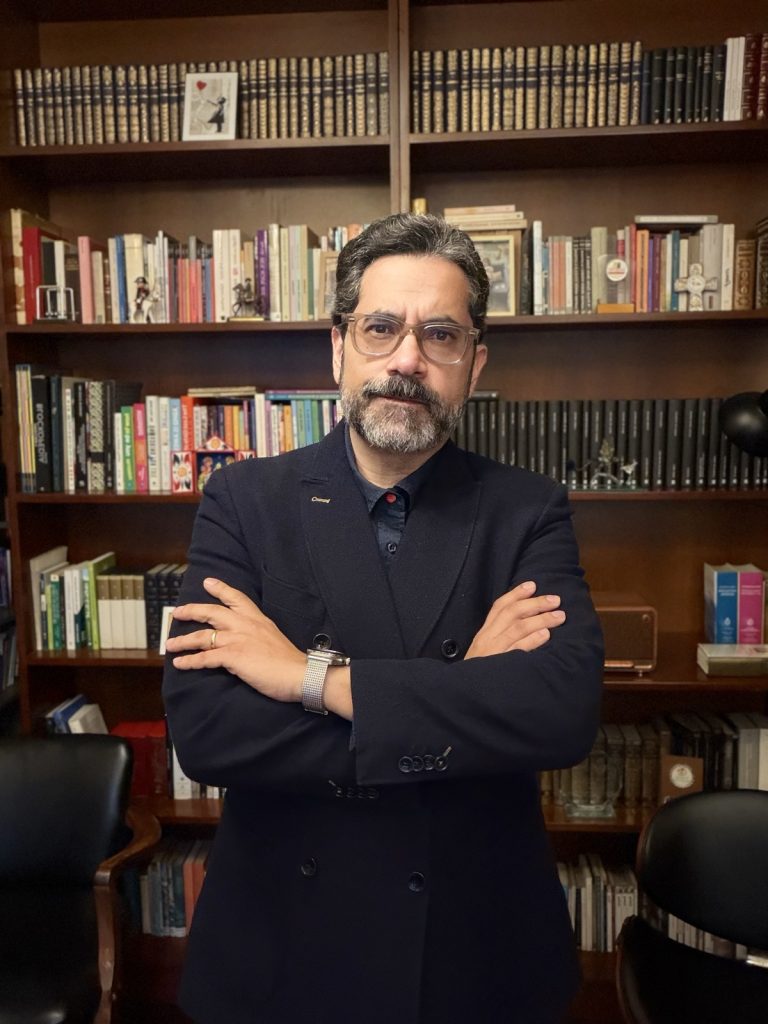 Ernesto Reaño de pie, con los brazos cruzados, frente a una biblioteca.